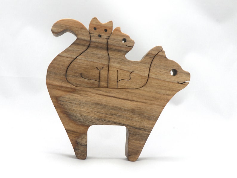 A freestanding wooden puzzle cut in the shape of a large cat standing on four legs. Two smaller kitten figures nest on the large cat’s back and interlock as puzzle pieces. Simple drilled holes form the eyes and nose on each cat, with a shallow cut line for the mouth on the large cat. The completed puzzle stands upright on a plain white surface. Handmade by Odin’s Toy Factory In Tallahassee, Florida, USA.