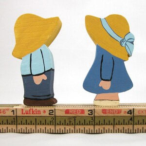May include: Two wooden figurines of people wearing yellow hats. The person on the left is wearing a blue shirt and blue pants. The person on the right is wearing a blue dress. Both figurines are standing on a wooden ruler marked with inches.