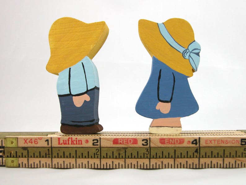 May include: Two wooden figurines of people wearing yellow hats. The person on the left is wearing a blue shirt and blue pants. The person on the right is wearing a blue dress. Both figurines are standing on a wooden ruler marked with inches.