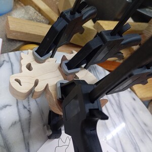A light wood dragon lies on a workbench while three clamps hold dark wood pieces in place during assembly.