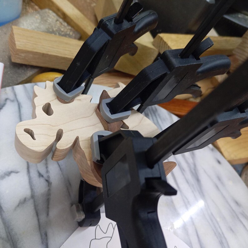 A light wood dragon lies on a workbench while three clamps hold dark wood pieces in place during assembly.