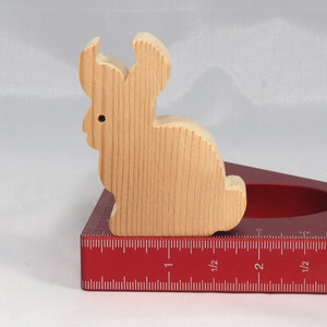 May include: A wooden rabbit figurine sits on a red protractor with inch markings. The rabbit is light brown and has a simple design.