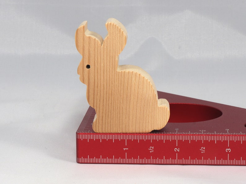May include: A wooden rabbit figurine sits on a red protractor with inch markings. The rabbit is light brown and has a simple design.