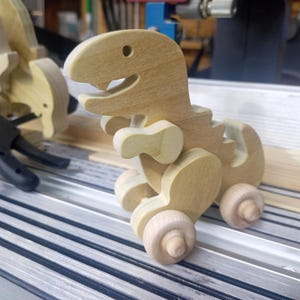 Wooden unfinished T-Rex dinosaur push toy on a ribbed metal Shopsmith table saw, showing its drilled eye, open mouth, short arms, curved tail, and wooden wheels, with clamps and another dinosaur behind it.