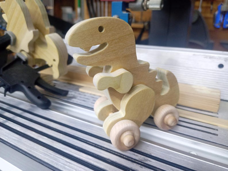 Wooden unfinished T-Rex dinosaur push toy on a ribbed metal Shopsmith table saw, showing its drilled eye, open mouth, short arms, curved tail, and wooden wheels, with clamps and another dinosaur behind it.