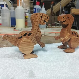 Two handmade wooden baby dinosaur toys standing on a toymaker’s workbench, showing their hardwood construction, separate wooden arms and legs, and three-dimensional shape.
