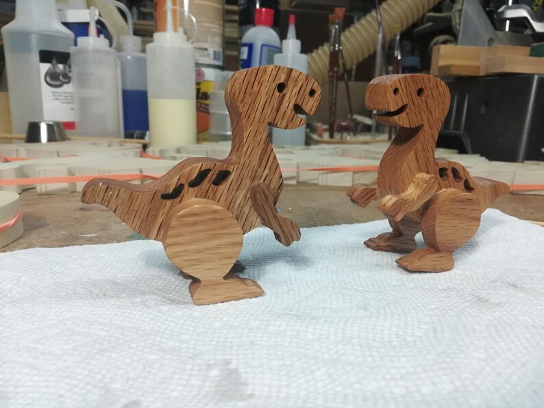 Two handmade wooden baby dinosaur toys standing on a toymaker’s workbench, showing their hardwood construction, separate wooden arms and legs, and three-dimensional shape.