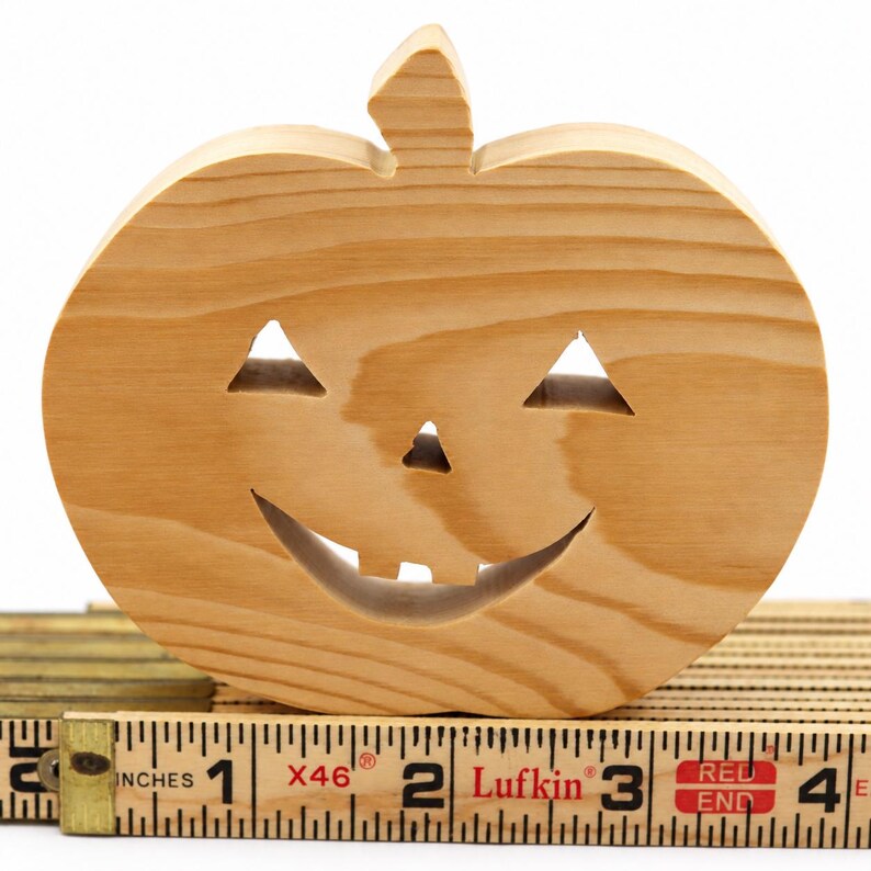 Handmade unpainted wooden Jack-o-Lantern cutout with carved triangular eyes, nose, smiling mouth, and stem, shown freestanding on a white background. Ready to paint for Halloween decoration. Sitting on a carpenter's rule for scale.