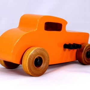 May include: A bright orange wooden toy car with a classic hot rod design. The car features wooden wheels, black exhaust pipes, and a glossy finish. The wheels are approximately 3 inches in diameter.