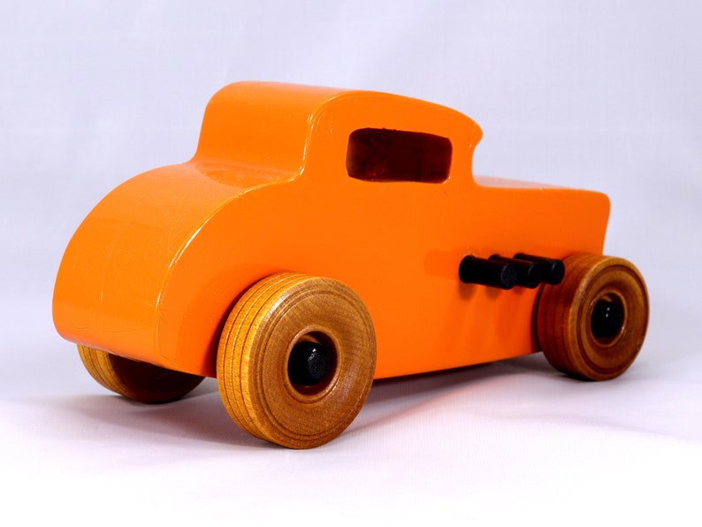 May include: A bright orange wooden toy car with a classic hot rod design. The car features wooden wheels, black exhaust pipes, and a glossy finish. The wheels are approximately 3 inches in diameter.