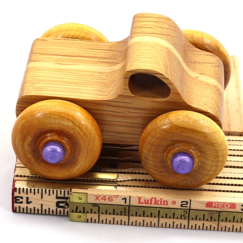 May include: A handcrafted wooden toy truck with four large, round wheels. The wheels have purple centers. The truck is made of light-colored wood and sits on a vintage measuring tape.