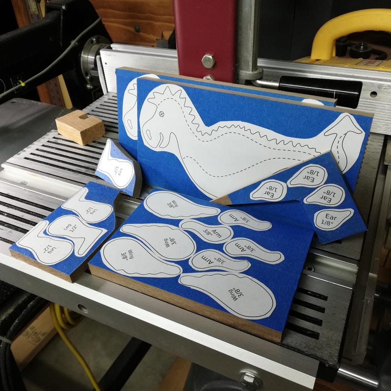 Baby dragon patterns attached to hardwood boards with blue painter’s tape, ready for cutting in the woodworking shop.