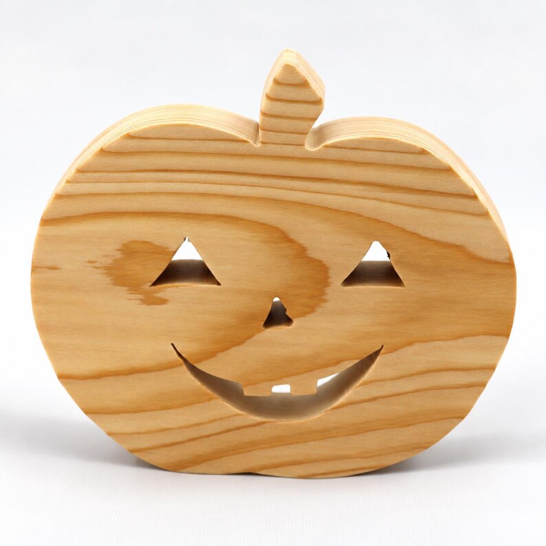 May include: A light brown wooden pumpkin cutout featuring a carved jack-o'-lantern face with two triangular eyes, a small triangular nose, and a wide smiling mouth with two square teeth. The natural wood grain is visible across the surface.