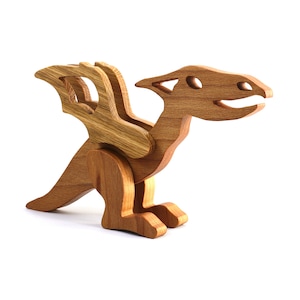 May include: A light brown wooden dragon figurine featuring a smooth finish, visible wood grain, a long tail, two upright wings, and a head with an open mouth. The creature is depicted in a standing position with two legs and a pointed snout.