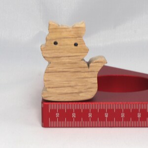 May include: A wooden cat figurine with black eyes, sitting on a red ruler with white markings. The ruler is marked in inches from 10 to 80.