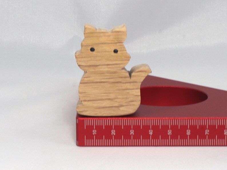 May include: A wooden cat figurine with black eyes, sitting on a red ruler with white markings. The ruler is marked in inches from 10 to 80.