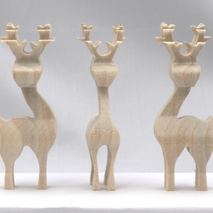 May include: Three wooden reindeer figurines. Each reindeer is carved from light-colored wood and features a slender neck, stylized antlers, and a simple body. The reindeer are standing on a white surface.