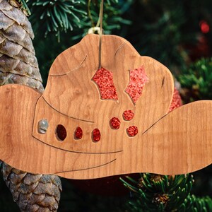 Handmade wooden Christmas tree ornament shaped like a cowboy hat, featuring a sprig of holly tucked into the hat band, star-shaped leaf cutouts, and round berry accents.