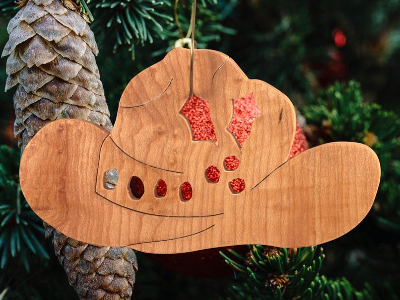 Handmade wooden Christmas tree ornament shaped like a cowboy hat, featuring a sprig of holly tucked into the hat band, star-shaped leaf cutouts, and round berry accents.