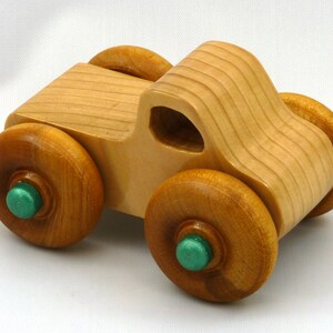 A handmade wooden toy monster truck with a natural wood body and four large wheels. The body and wheels are finished with amber shellac, and the hubs are painted with metallac emerald green acrylic paint. The toy is designed with a simple, classic aesthetic.