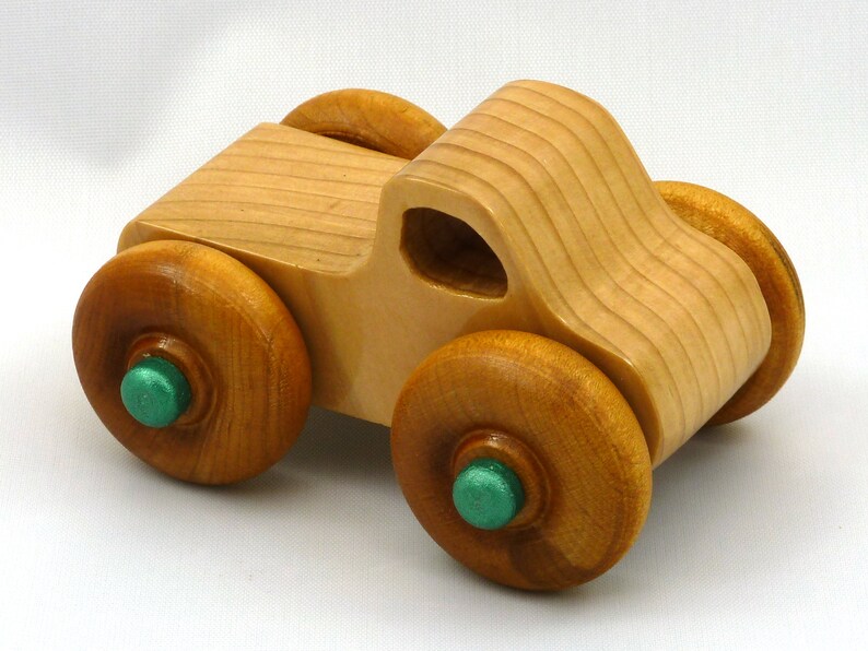 A handmade wooden toy monster truck with a natural wood body and four large wheels. The body and wheels are finished with amber shellac, and the hubs are painted with metallac emerald green acrylic paint. The toy is designed with a simple, classic aesthetic.