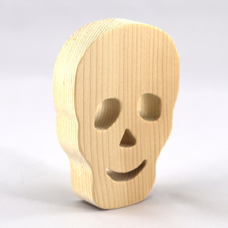 A handcrafted, unfinished light wood Halloween skull cutout featuring carefully carved eye sockets, a nose, and a cheerful, smiling mouth. Displayed upright against a white background.