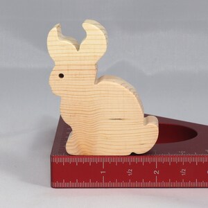 May include: A wooden rabbit figurine, painted in a natural wood color, is sitting on a red measuring tool with a ruler marked in inches. The rabbit has long ears and a rounded body.