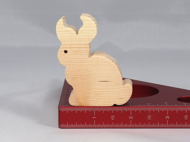 May include: A wooden rabbit figurine, painted in a natural wood color, is sitting on a red measuring tool with a ruler marked in inches. The rabbit has long ears and a rounded body.