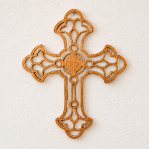 Handmade wooden cross with warm amber wood grain, intricate Victorian-style fretwork, and a round center, hanging from a small nail on a lightly textured off-white wall.
