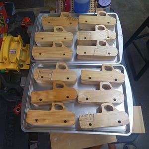 Unfinished wooden toy pickup bodies arranged on two metal trays, with cab windows cut out and axle holes drilled.