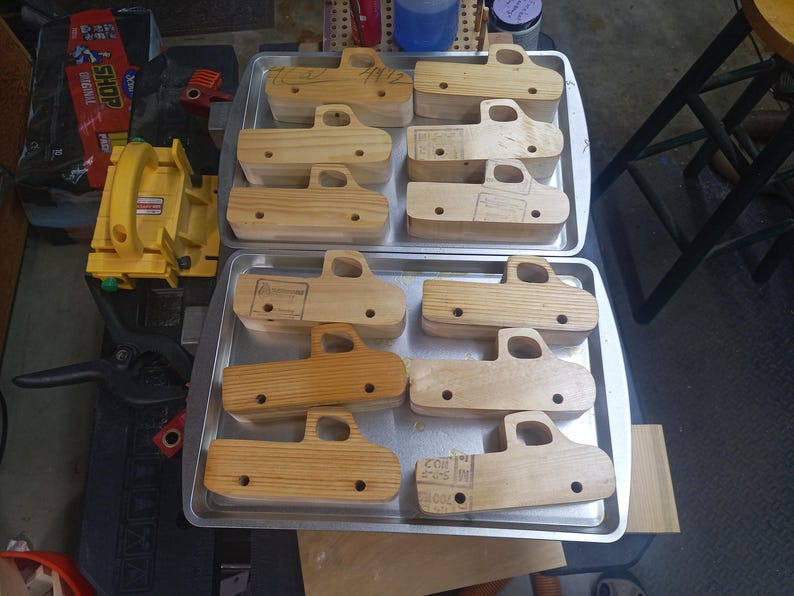 Unfinished wooden toy pickup bodies arranged on two metal trays, with cab windows cut out and axle holes drilled.