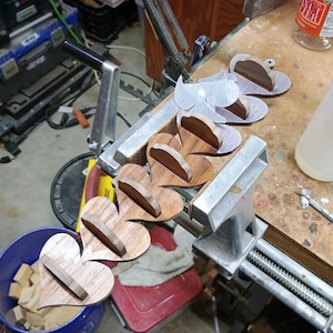 May include: A series of connected heart-shaped wooden pieces held in a metal vise. Each heart features a small, perpendicular wooden shelf attached to its center. The wood displays a natural brown grain pattern, and the pieces are arranged in a line.