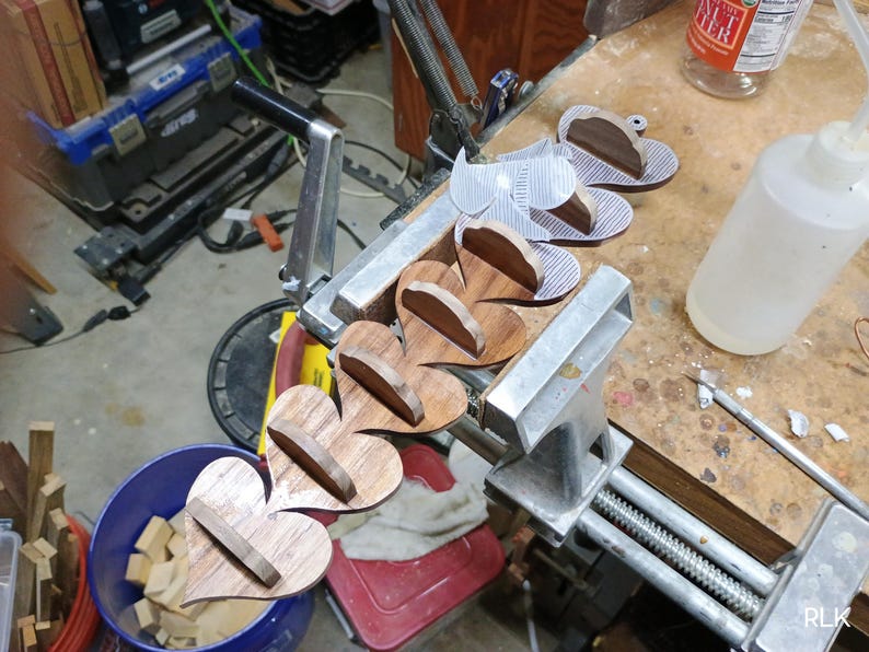 May include: A series of connected heart-shaped wooden pieces held in a metal vise. Each heart features a small, perpendicular wooden shelf attached to its center. The wood displays a natural brown grain pattern, and the pieces are arranged in a line.
