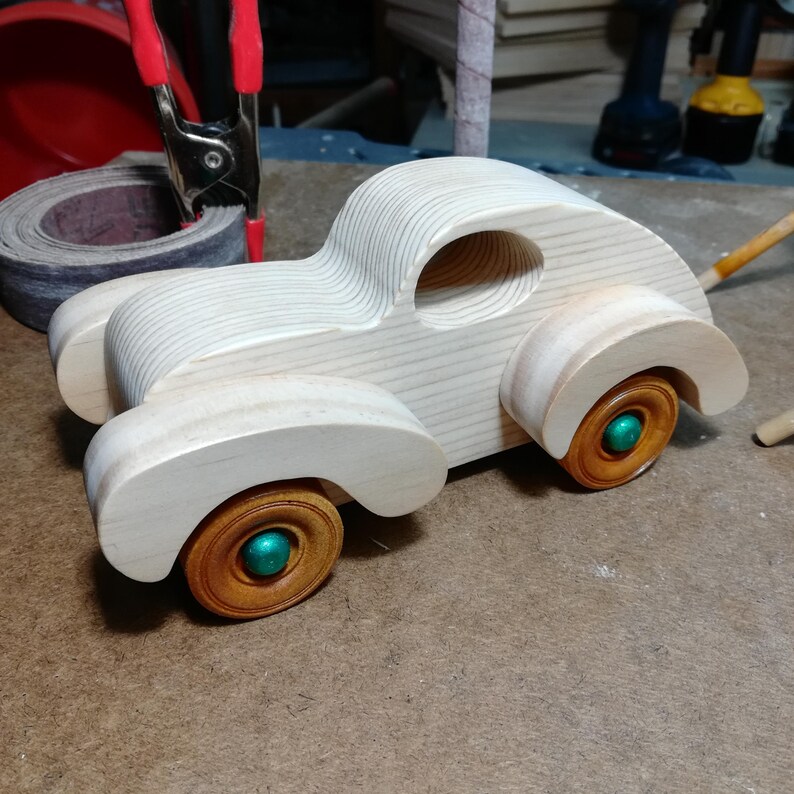 An assembled wooden fat-fender toy car rests on a workshop bench with its front pointing left. The unpainted pine body shows distinct grain lines across the long hood, curved roof, and oval window opening. Broad, sanded fenders arch over amber-brown wooden wheels with metallic green axle ends. A rolled sanding belt held by a red-handled spring clamp sits behind the car, with tools and stacked wood in the background.