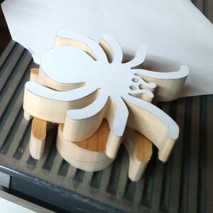 Stack of handmade unfinished wooden spider cutouts in my workshop, showing their 3/4-inch thickness and layered wood grain. A white paper pattern is attached to the top spider. These unpainted, freestanding Halloween decorations are hand-cut from solid wood and ready for painting or crafting.