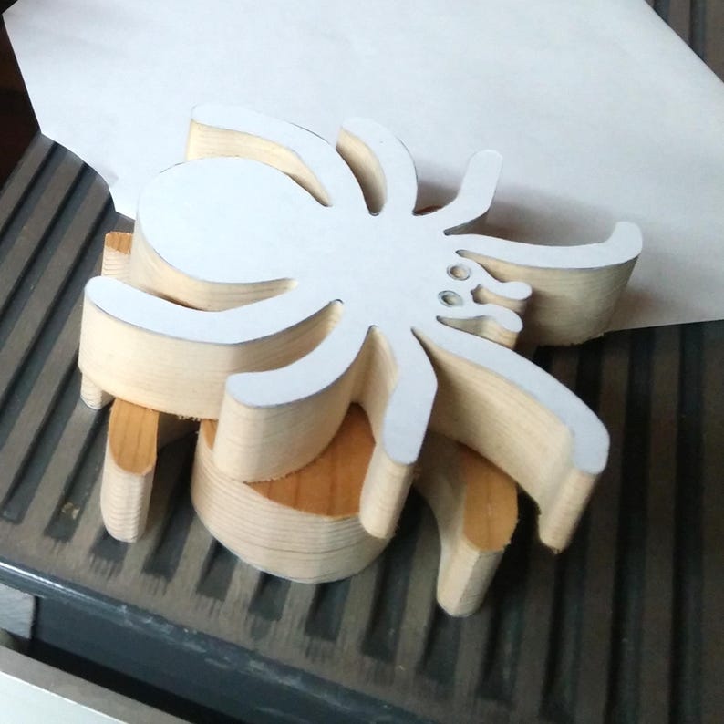 Stack of handmade unfinished wooden spider cutouts in my workshop, showing their 3/4-inch thickness and layered wood grain. A white paper pattern is attached to the top spider. These unpainted, freestanding Halloween decorations are hand-cut from solid wood and ready for painting or crafting.