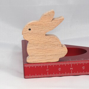 May include: A wooden rabbit figurine sits on a red, triangular ruler with inch markings. The ruler has a cut-out circle in the center.