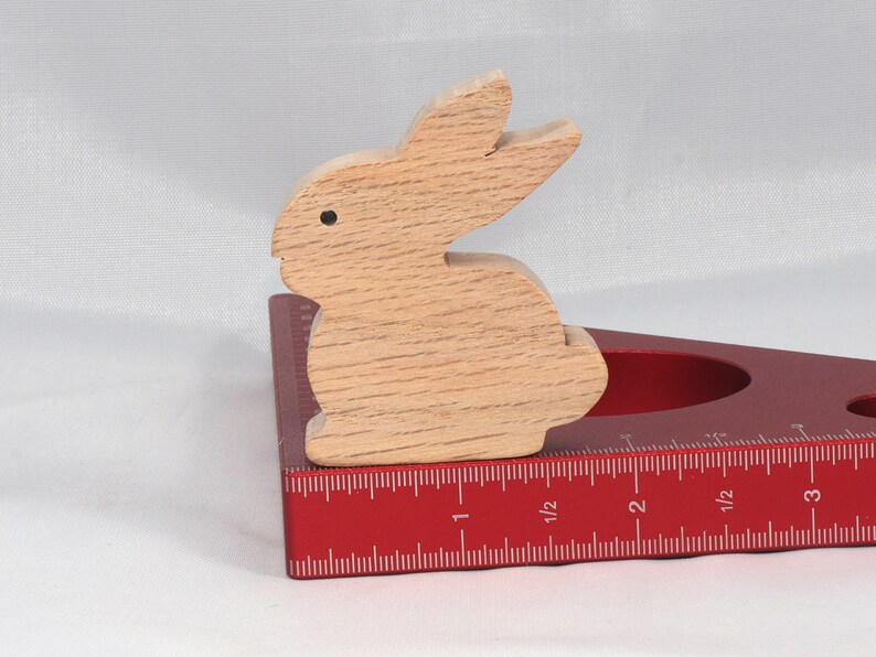 May include: A wooden rabbit figurine sits on a red, triangular ruler with inch markings. The ruler has a cut-out circle in the center.