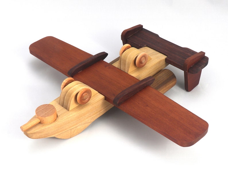 May include: A handcrafted wooden toy airplane with a biplane design. The wings and tail are a rich, dark brown, contrasting with the lighter natural wood of the fuselage and wheels. The propeller is also made of light wood.