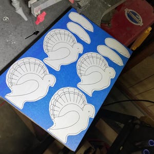 Paper turkey and wing patterns arranged on wood covered with blue painter’s tape beside a scroll saw blade in the workshop.