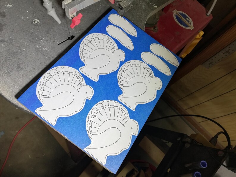 Paper turkey and wing patterns arranged on wood covered with blue painter’s tape beside a scroll saw blade in the workshop.