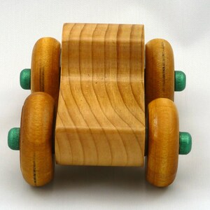 A handmade wooden toy monster truck with a natural wood body and four large wheels. The body and wheels are finished with amber shellac, and the hubs are painted with metallac emerald green acrylic paint. The toy is designed with a simple, classic aesthetic.