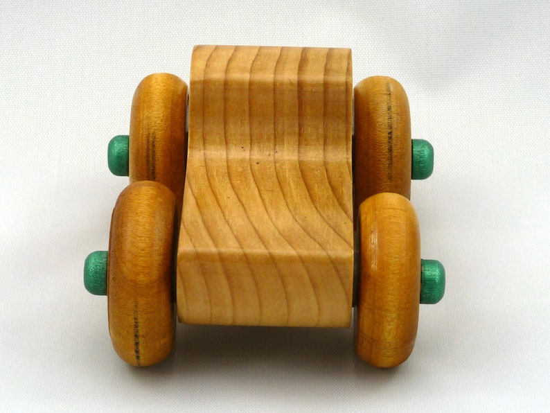 A handmade wooden toy monster truck with a natural wood body and four large wheels. The body and wheels are finished with amber shellac, and the hubs are painted with metallac emerald green acrylic paint. The toy is designed with a simple, classic aesthetic.