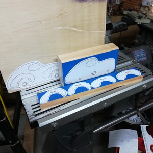 Wooden toy car patterns arranged on a Shopsmith bandsaw table in the workshop. A thick pine block holds the body pattern over blue painter’s tape, showing the oval window and axle-hole locations. A narrow board in front holds four curved fat fender patterns labeled “Rear” and “Front.” A full side-view drawing of the assembled car leans against a large wood panel behind the uncut pieces.