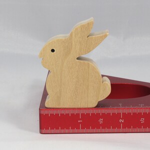 May include: A wooden rabbit cutout sits on a red measuring tool with a ruler marked in inches. The rabbit is light brown and has a simple design.