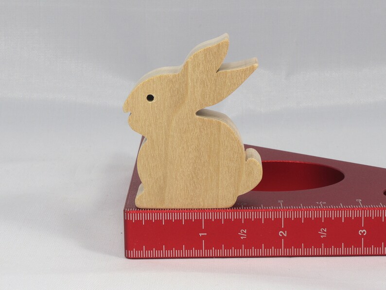 May include: A wooden rabbit cutout sits on a red measuring tool with a ruler marked in inches. The rabbit is light brown and has a simple design.