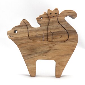 A freestanding wooden puzzle cut in the shape of a large cat standing on four legs. Two smaller kitten figures nest on the large cat’s back and interlock as puzzle pieces. Simple drilled holes form the eyes and nose on each cat, with a shallow cut line for the mouth on the large cat. The completed puzzle stands upright on a plain white surface. Handmade by Odin’s Toy Factory In Tallahassee, Florida, USA.