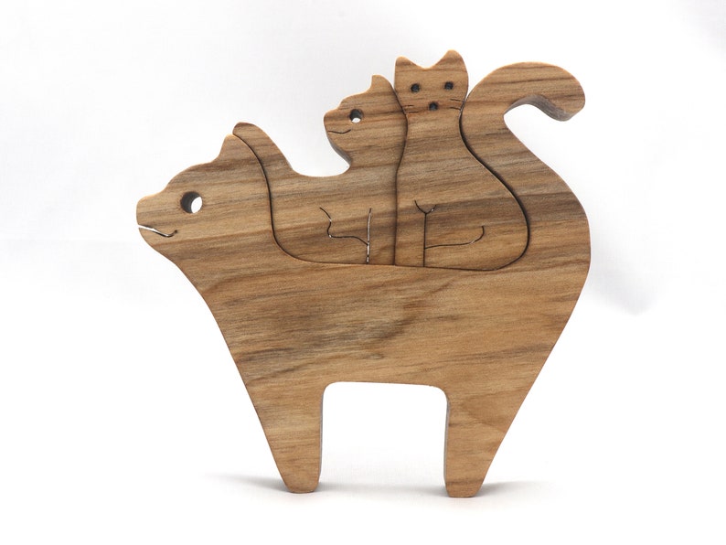 A freestanding wooden puzzle cut in the shape of a large cat standing on four legs. Two smaller kitten figures nest on the large cat’s back and interlock as puzzle pieces. Simple drilled holes form the eyes and nose on each cat, with a shallow cut line for the mouth on the large cat. The completed puzzle stands upright on a plain white surface. Handmade by Odin’s Toy Factory In Tallahassee, Florida, USA.