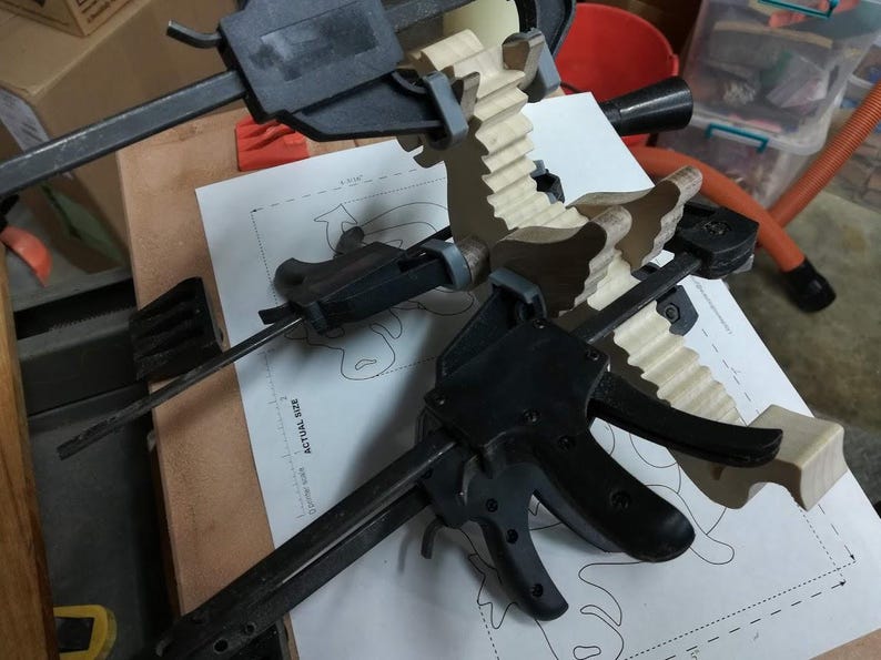 Wooden baby dragon figurine held with multiple clamps while its contrasting wood wings, ears, arms, and legs are glued in place.