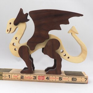 Handmade wooden dragon figurine made from contrasting hardwoods with a light natural wood body and head paired with rich dark reddish-brown wings, legs, and horns. Displayed on a wooden carpenters rule for scale.